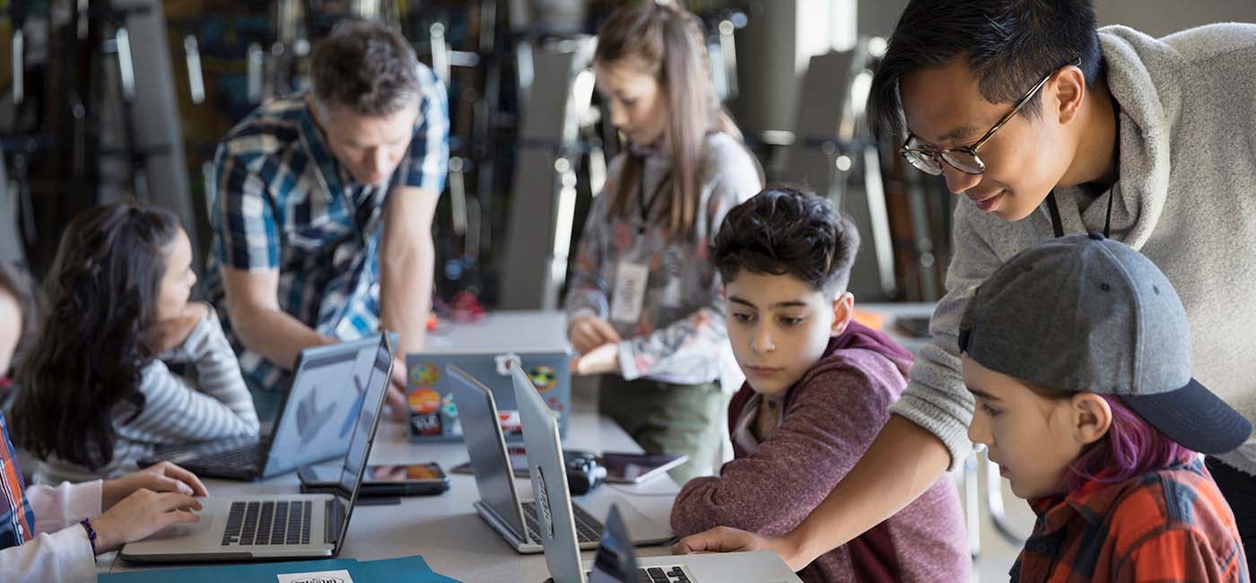 A photo of five children using laptops, sitting at a rectangle table in a classroom,  with two male teachers helping them