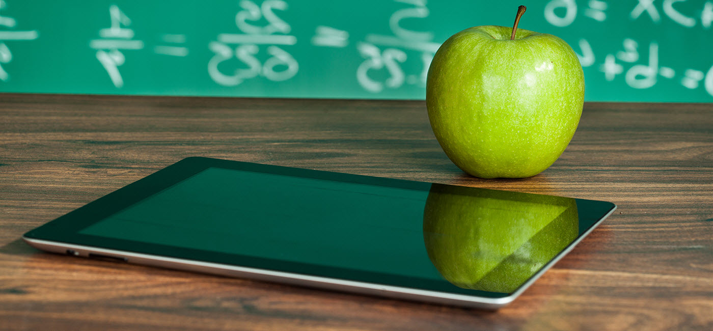 Ipad on a desk with an apple and chalkboard in the background.!''